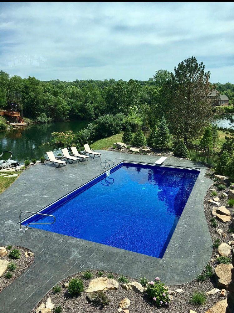 A large blue swimming pool surrounded by lawn chairs and a lake.