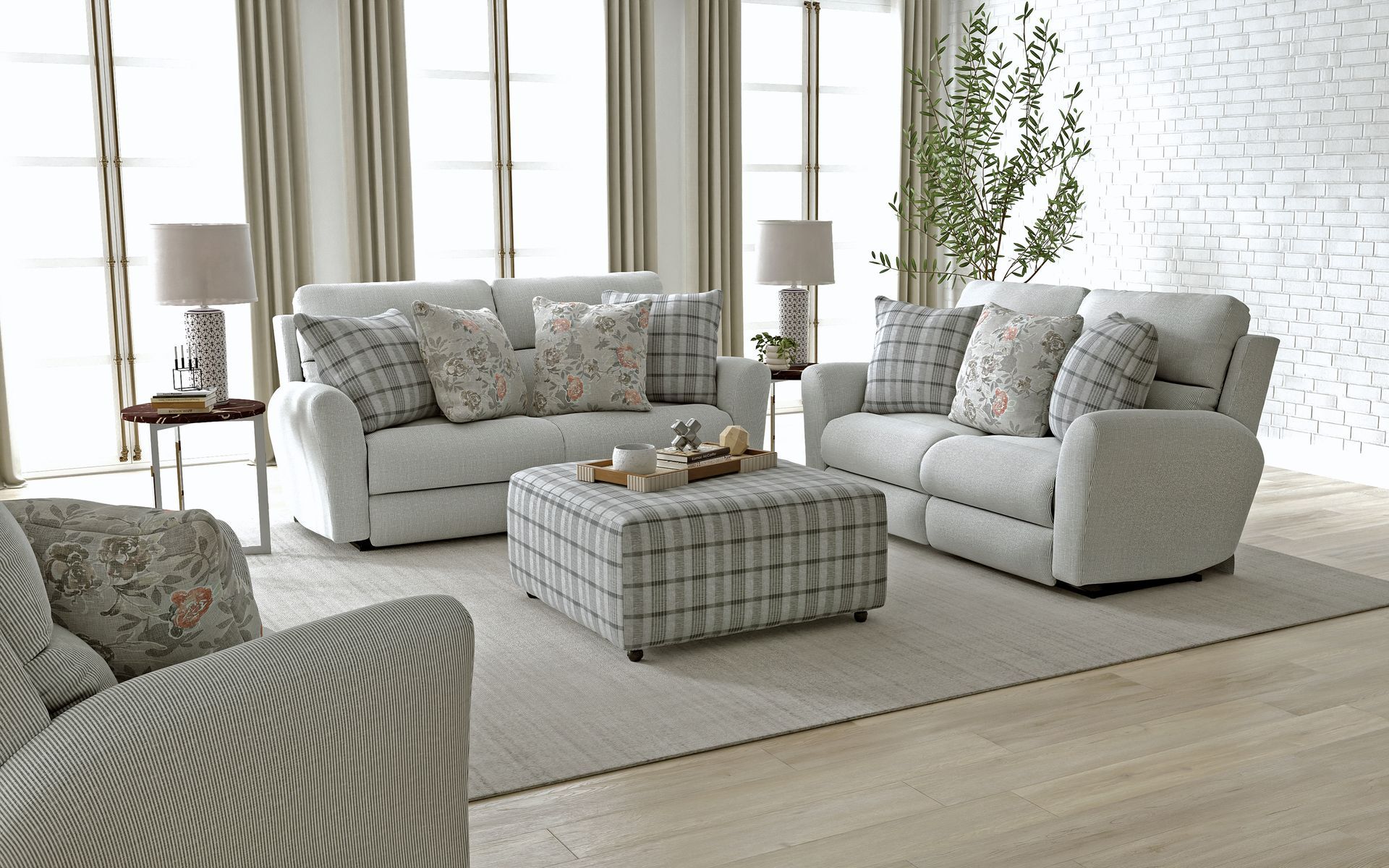 Bright living room with white sofas, patterned ottoman, lamps, and a light rug in front of a brick wall