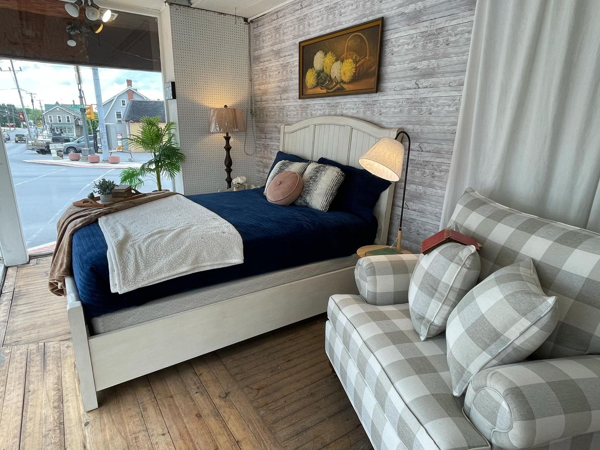 Bright bedroom showroom with navy bed, plaid armchair, and city view through large windows