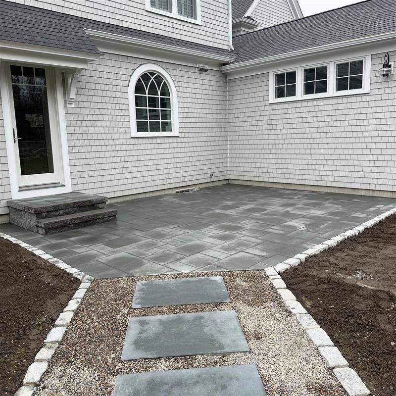 Exterior patio with gray pavers and stepping stones leading to a house with gray shingles.