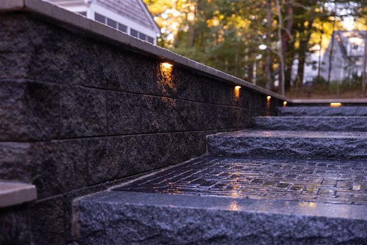 Stone steps with built-in lights along a dark stone wall, leading to a house with trees in the background.