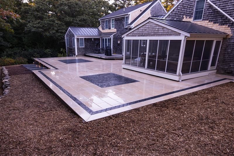 Stone patio with black border, adjacent to house and screened porch. Brown gravel ground.