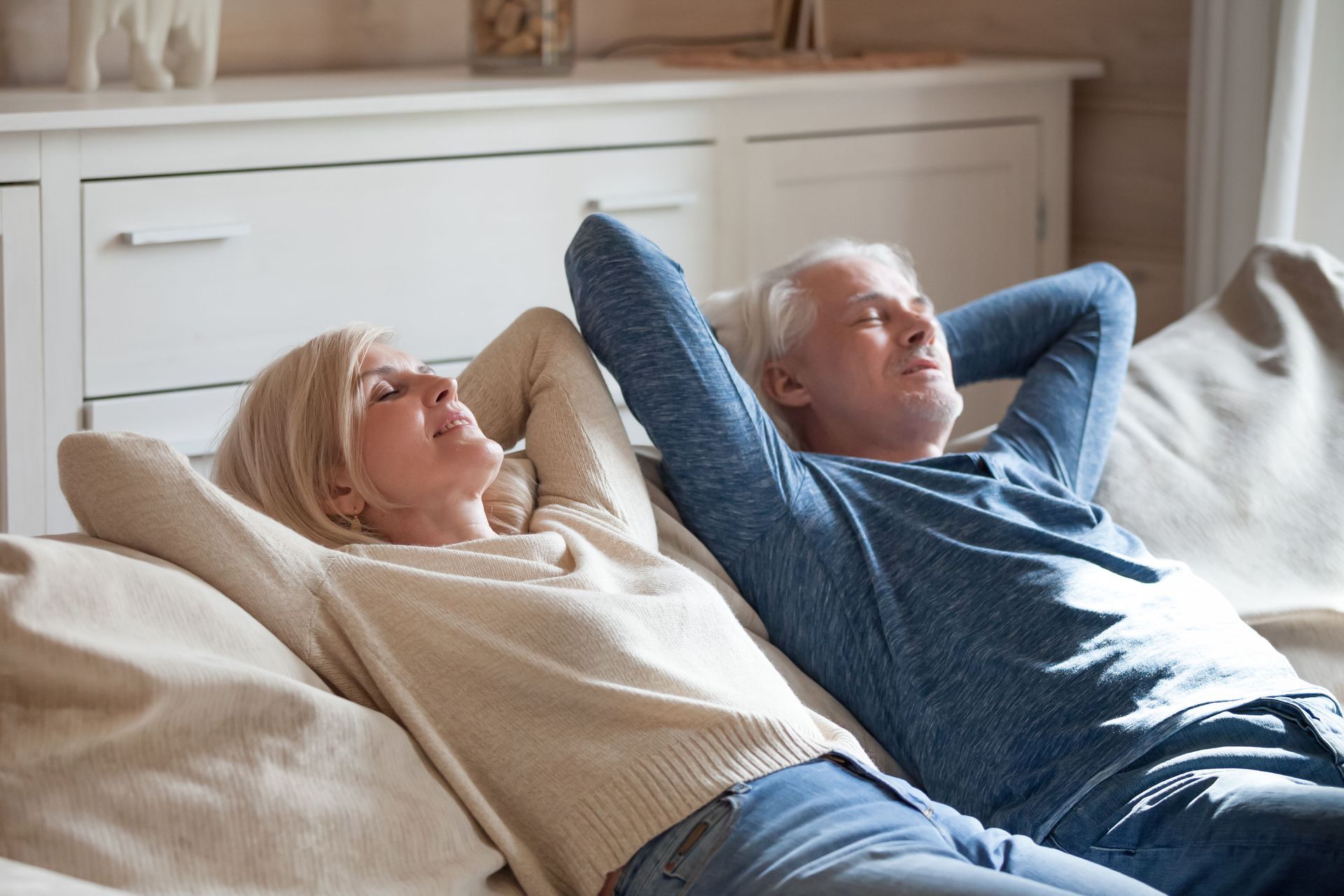An older couple relaxing on a couch indoors with their eyes closed, arms behind heads.
