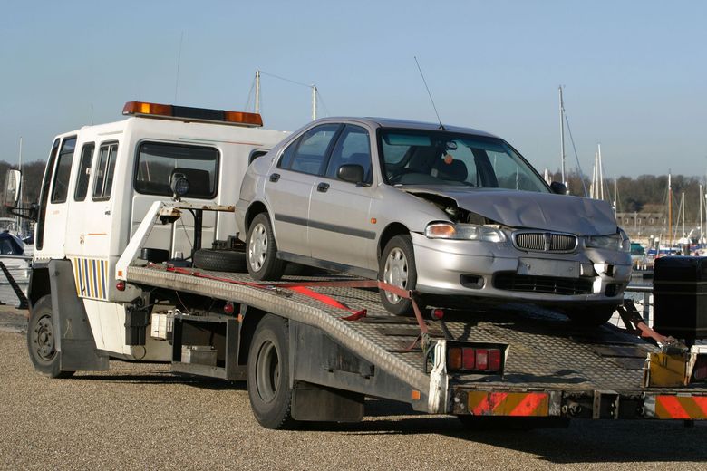 Tow truck carrying a damaged silver car, likely after a collision, in a harbor setting.