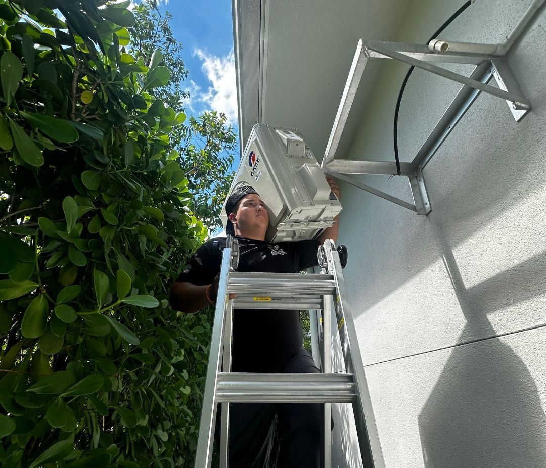 A man is standing on a ladder fixing an air conditioner on the side of a building.
