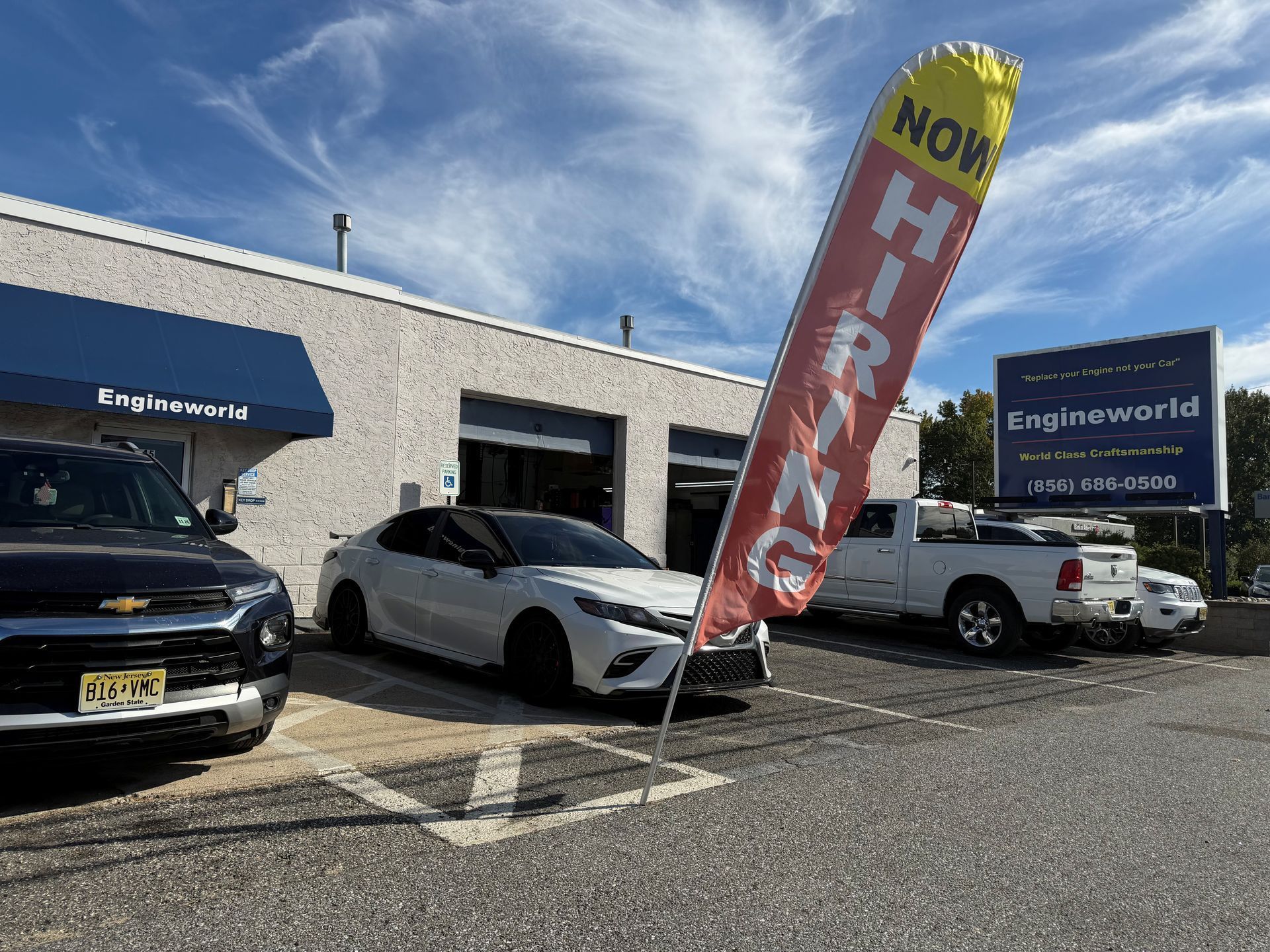Car dealership lot with cars parked beside a red “NOW HIRING” banner and building signage.