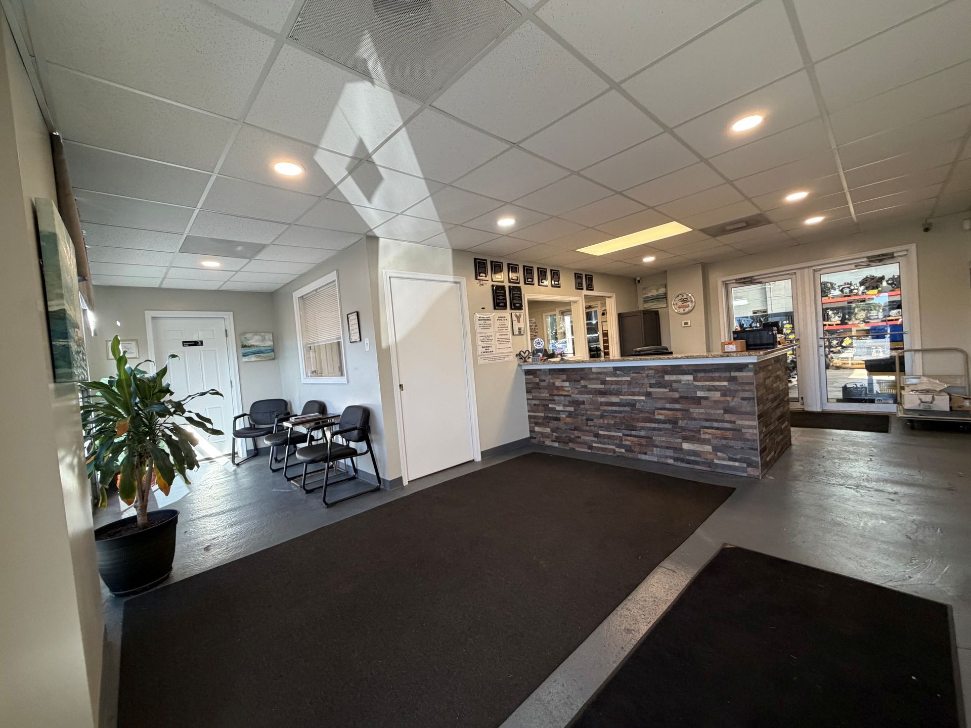 Bright office lobby with waiting chairs, a front desk, potted plant, and black floor mats