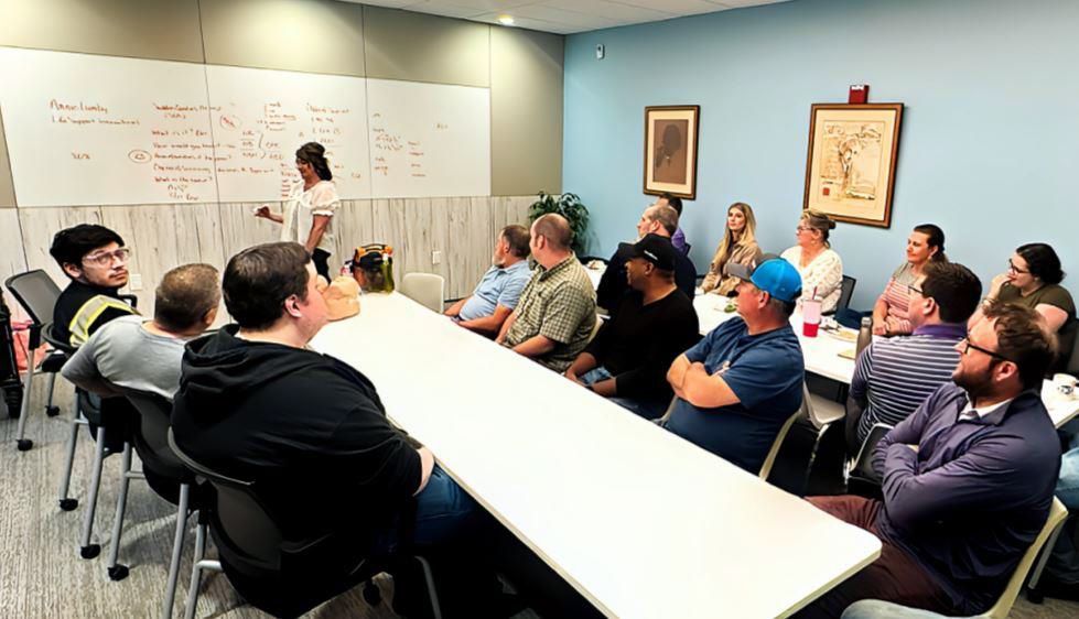 Team meeting in a conference room, with people seated around a white table as a presenter writes on a whiteboard.