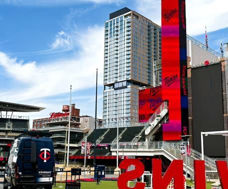 Downtown stadium scene with a tall tower, red sign, and a Minnesota Twins van in front
