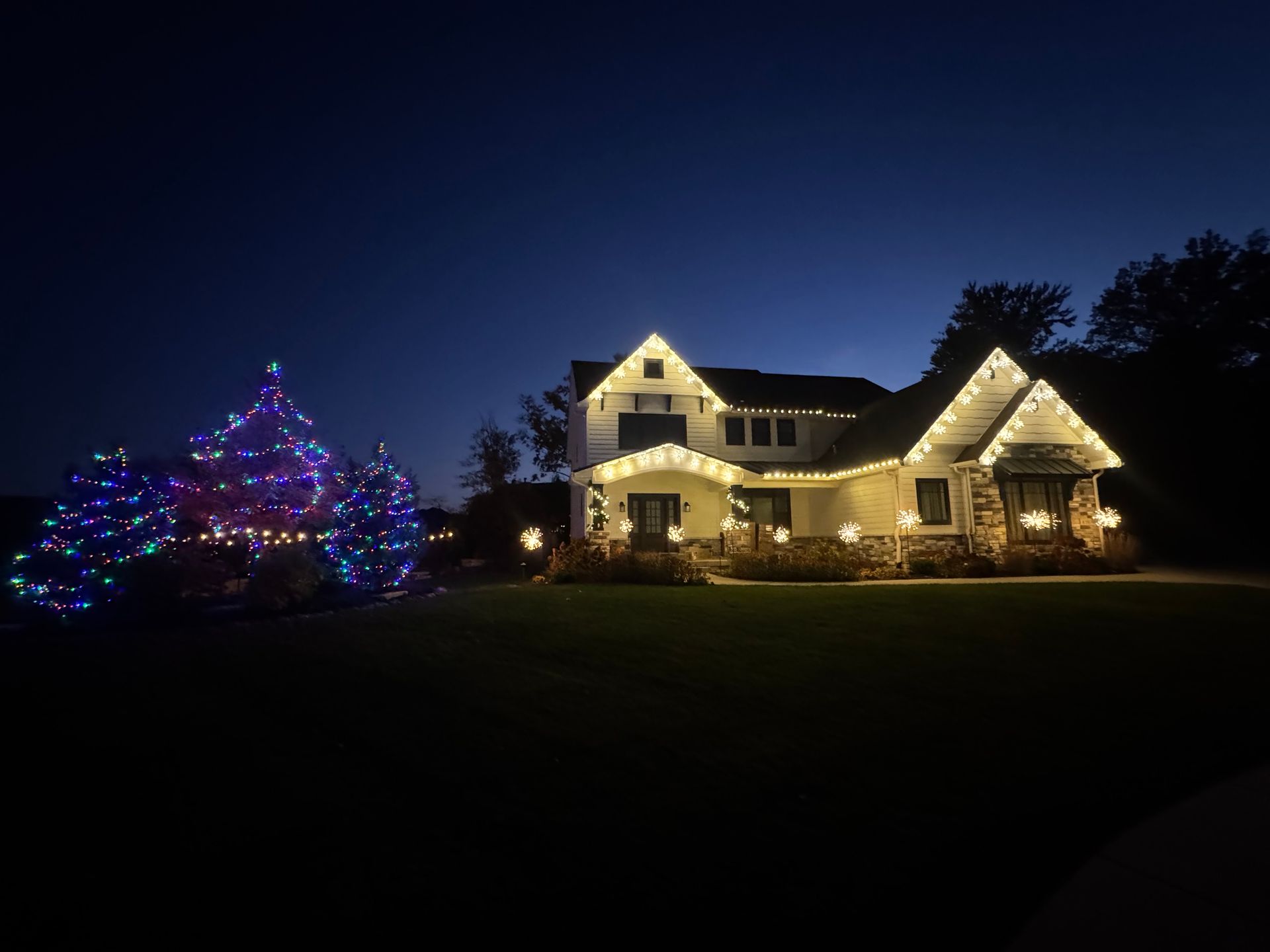 House and landscaping illuminated with colorful Christmas lights against a dark blue sky.