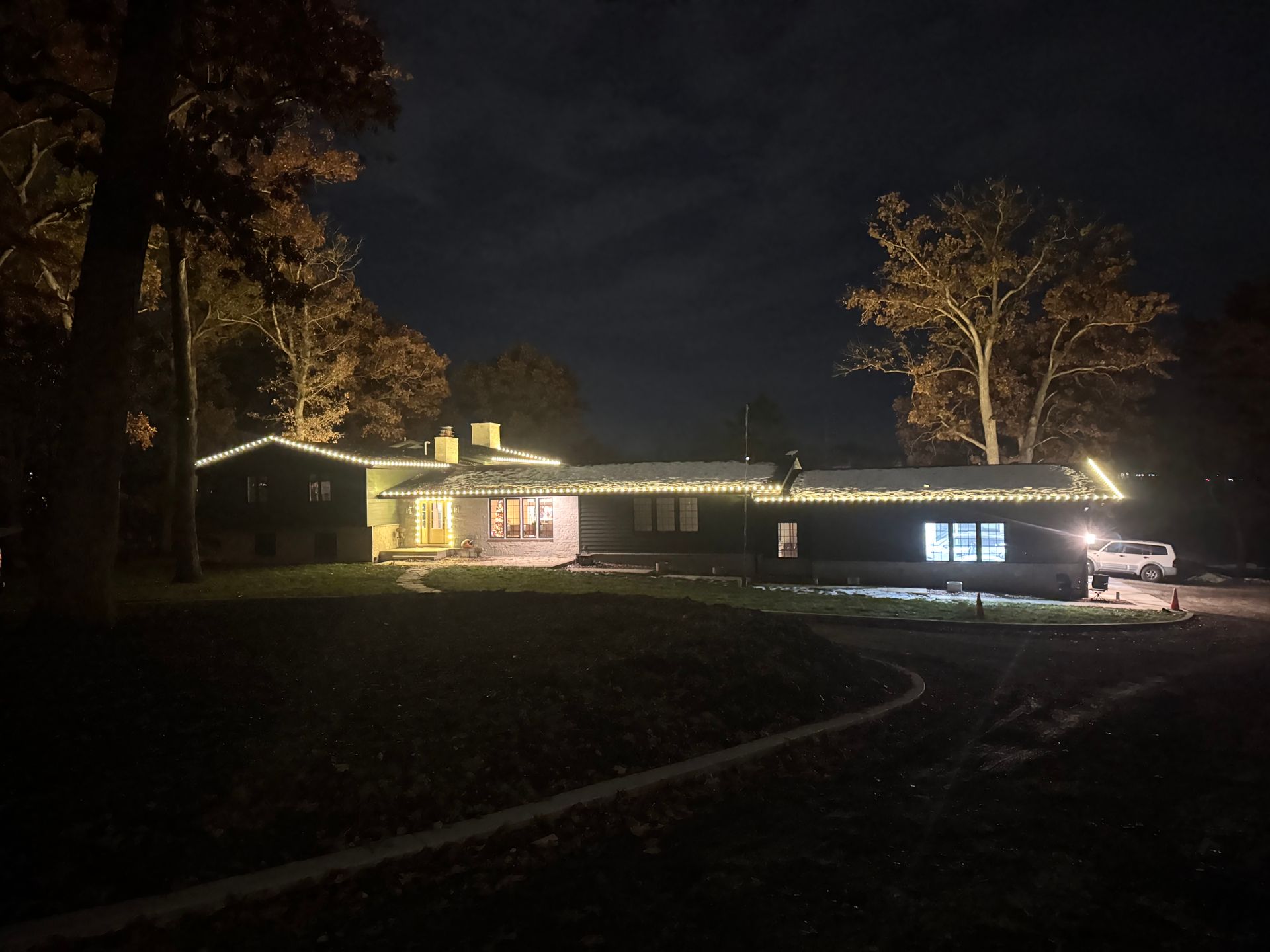 House at night with Christmas lights outlining roof and porch, car parked in driveway.