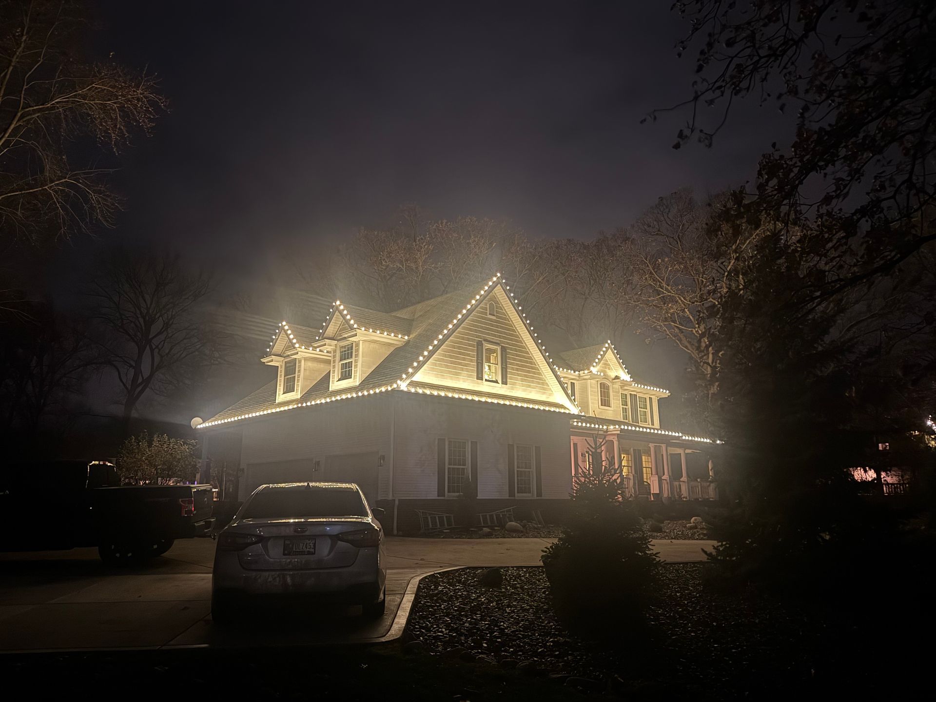 House at night with white lights along roof and porch; car parked in front.