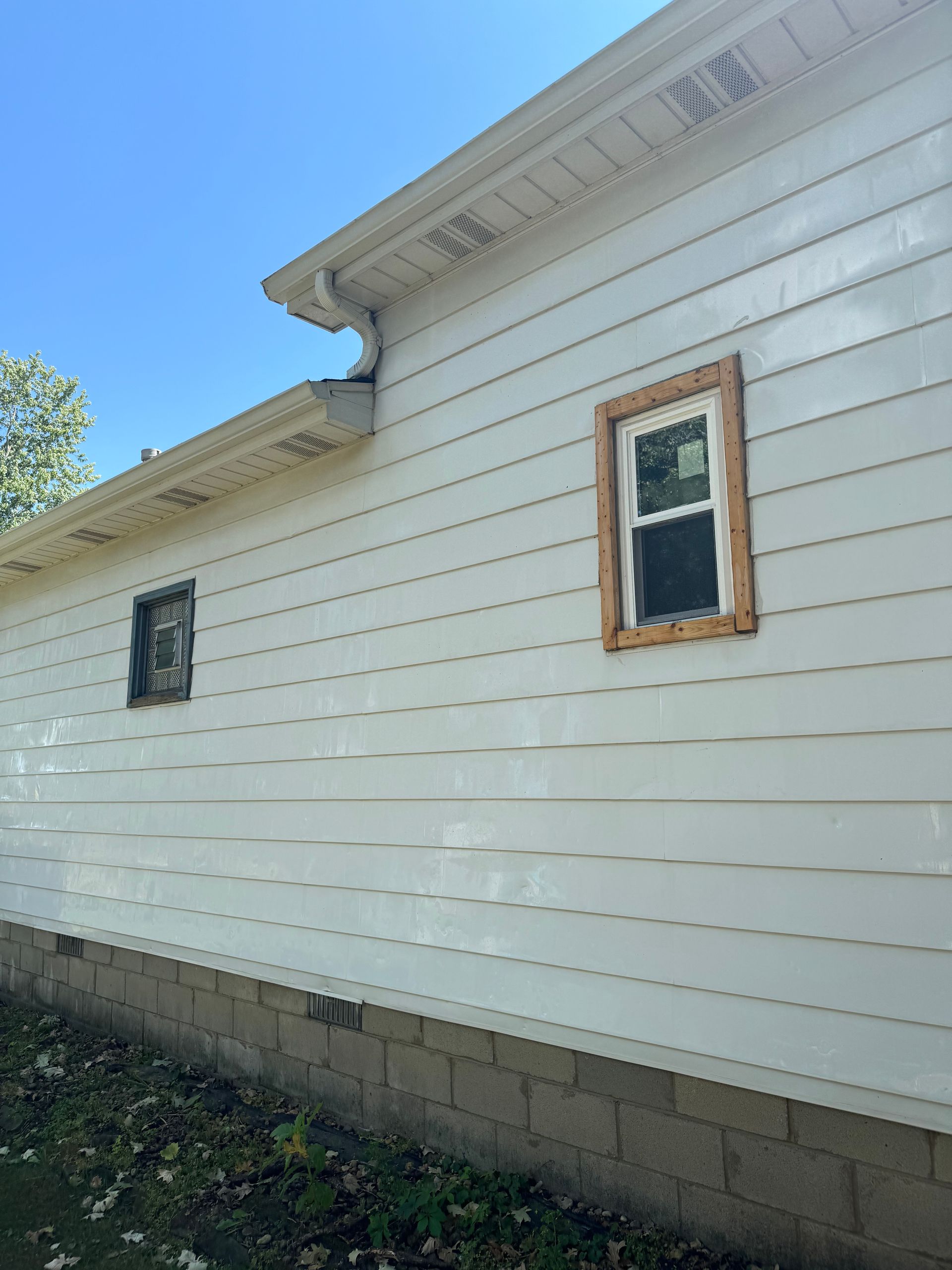 White house exterior with two windows and a tan wood trim. Brick foundation and gutter under a blue sky.
