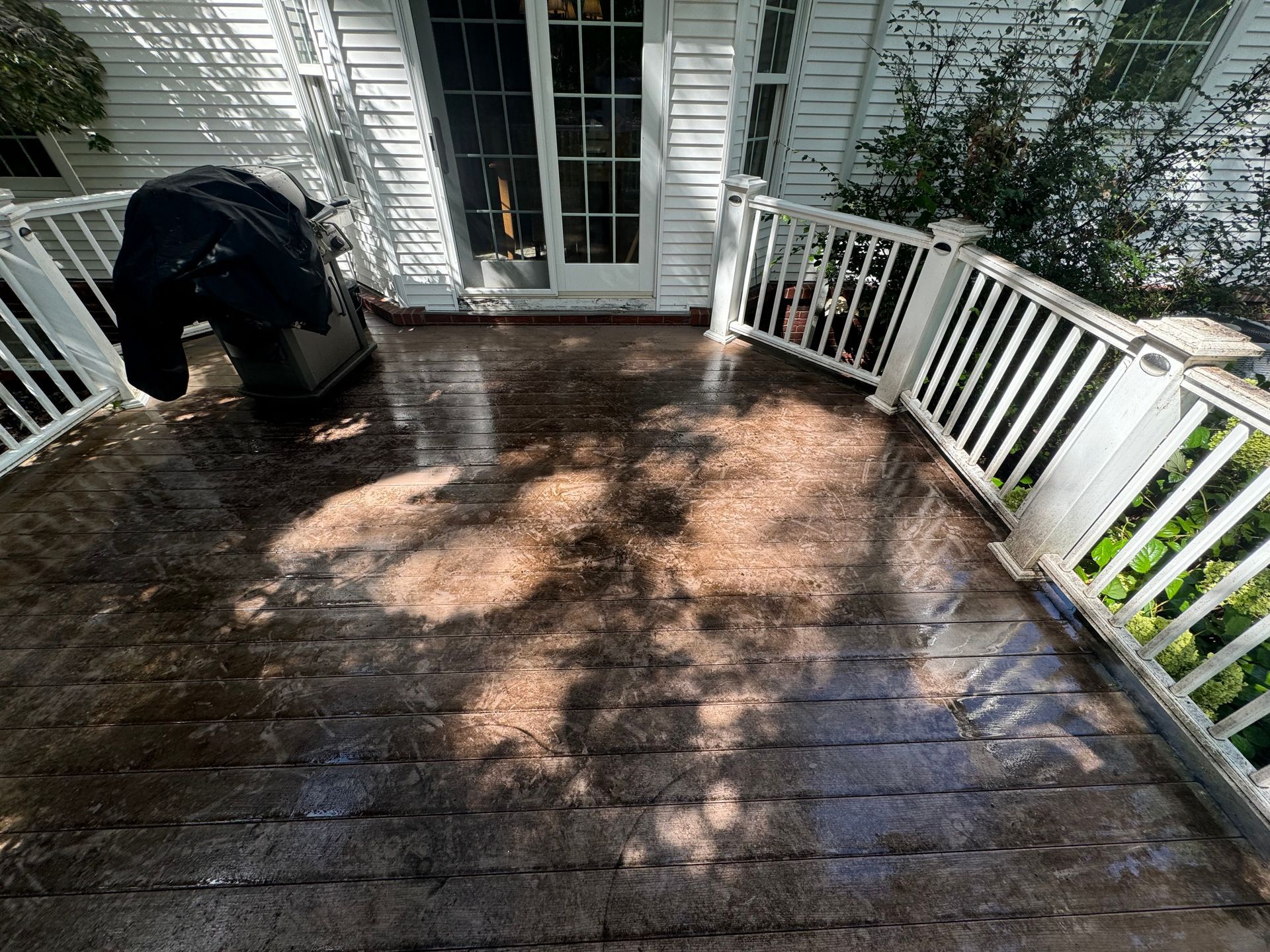 A person pressure washing a wooden deck with white railing next to a house with a glass door.