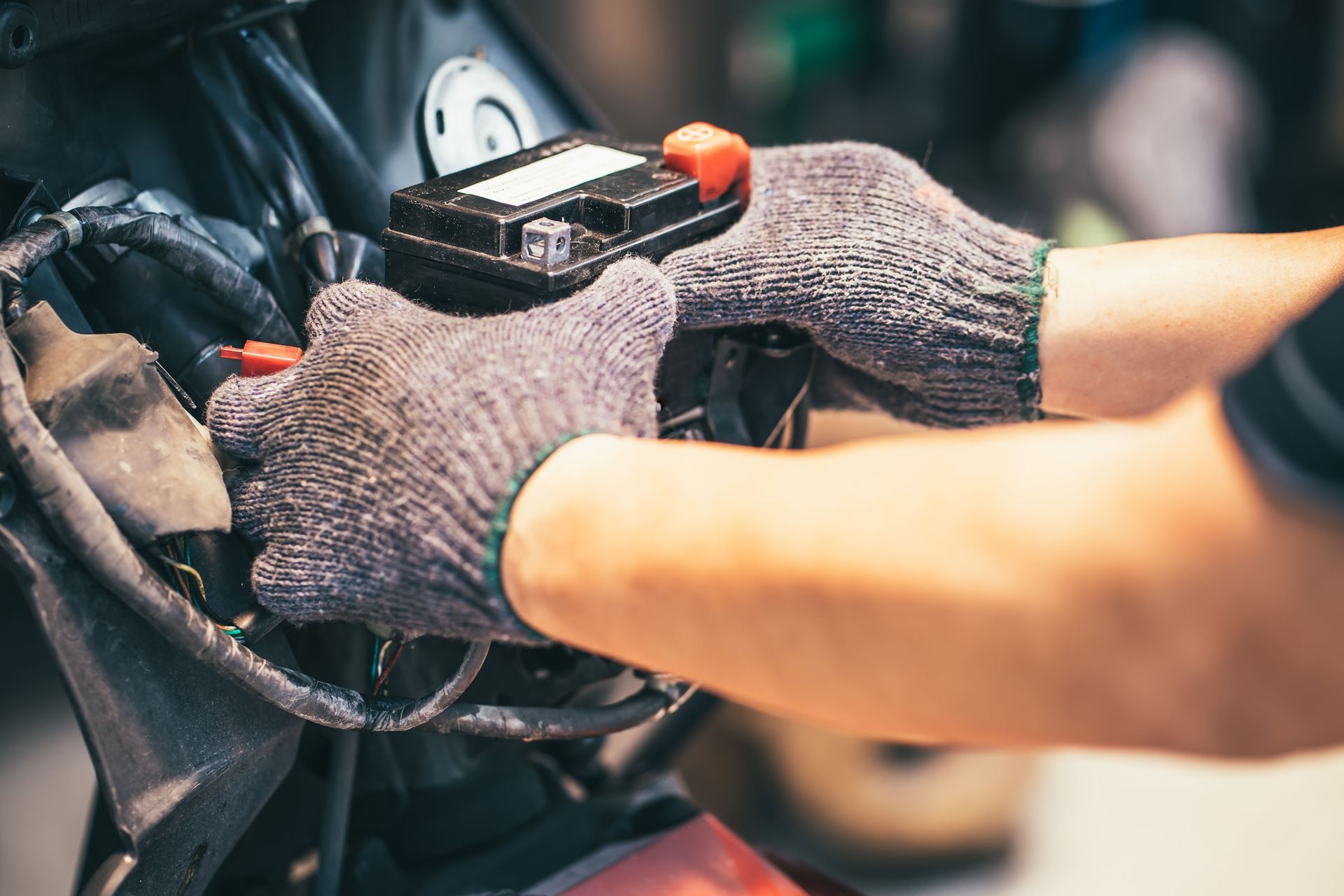 Mechanic wearing gray gloves installs or removes a small motorcycle battery from the engine compartment.