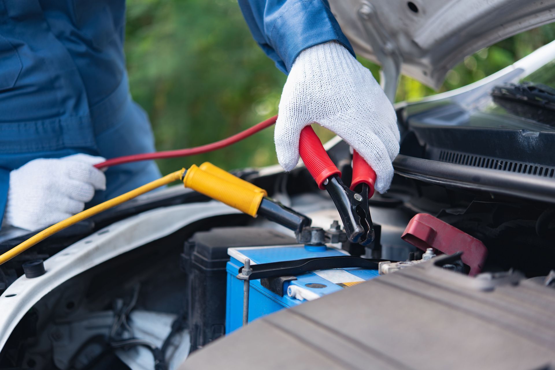 Mechanic wearing white gloves attaches jumper cables to a car battery under the hood.