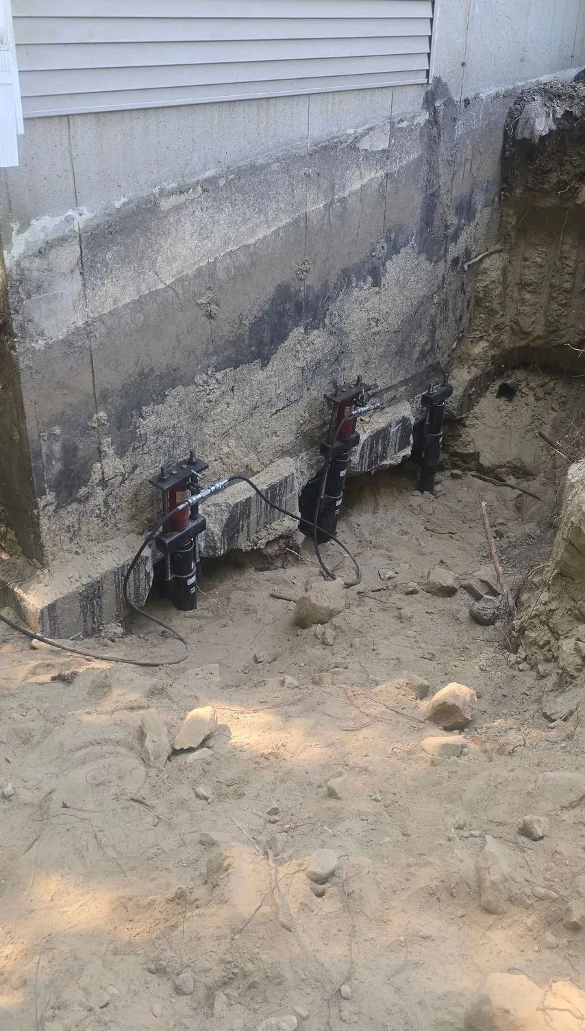 Damaged house foundation with exposed concrete, dirt excavation, and small utility pipes near the wall