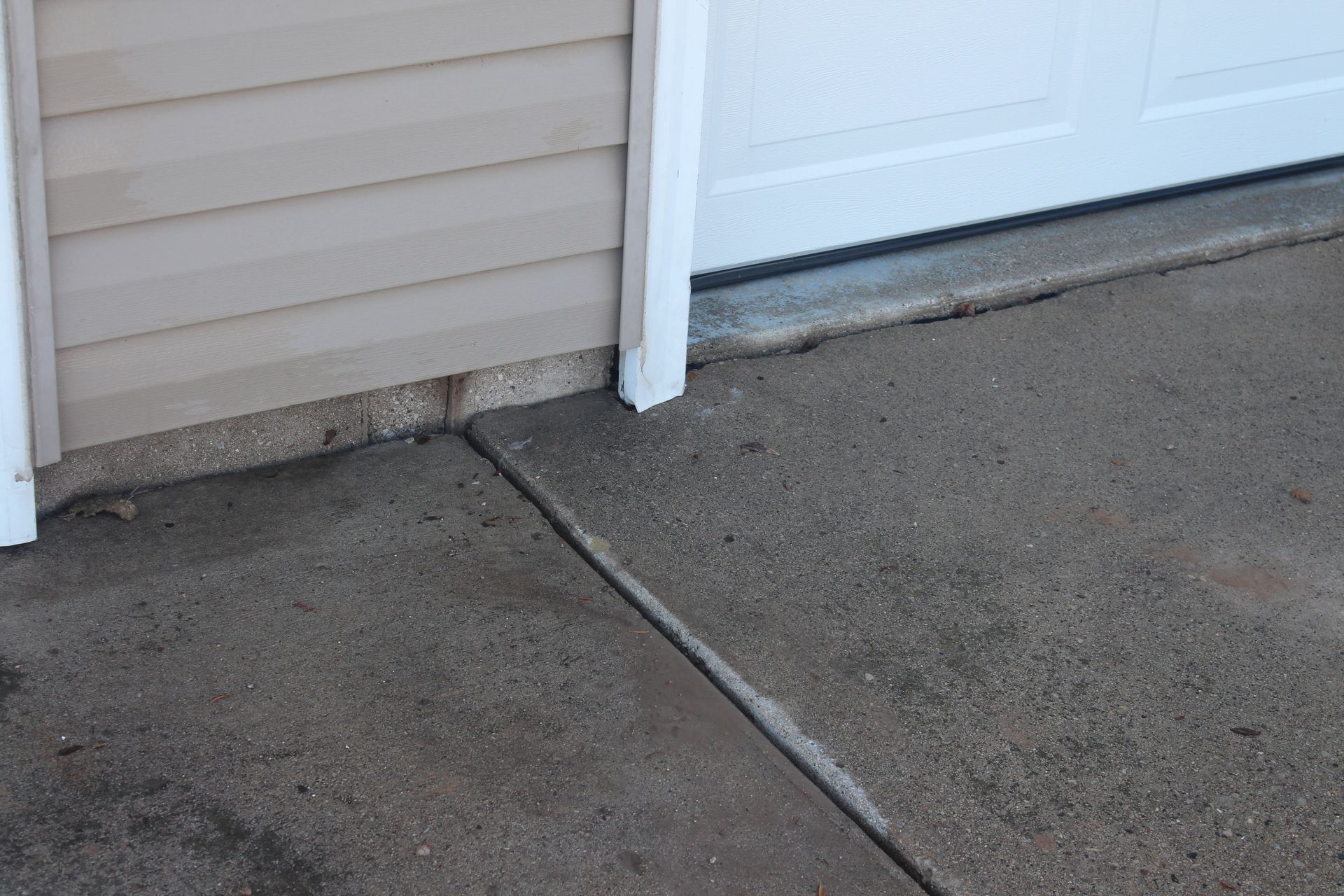 A gap between a concrete driveway and the foundation of a house with tan siding.