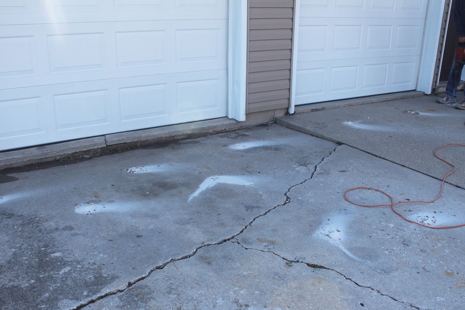 A cracked concrete driveway featuring white spray-painted markings indicating areas for repair near two garage doors.