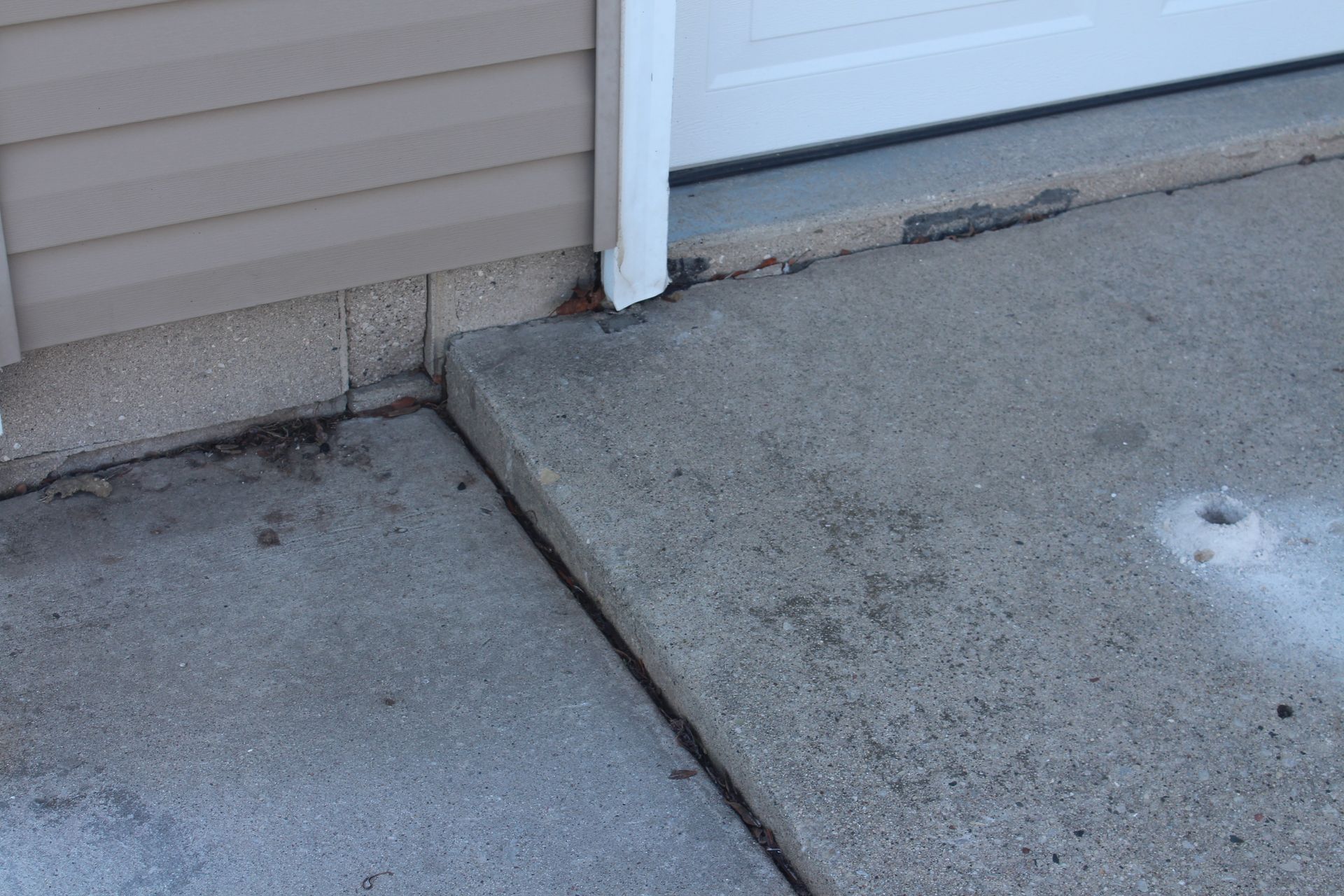 A concrete driveway edge showing a gap and height difference where it meets the side of a house and garage door.