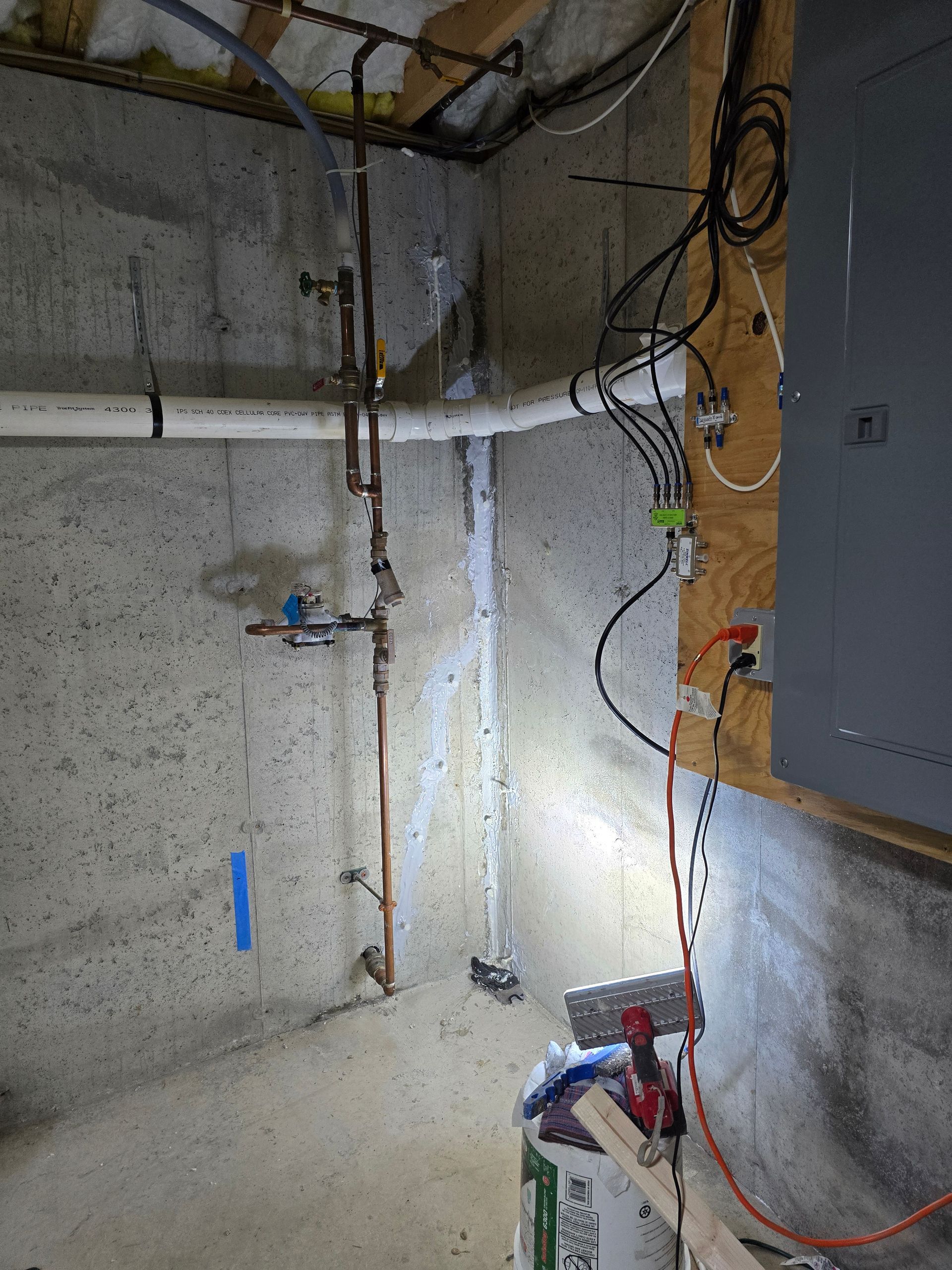 A concrete corner in a basement showing copper plumbing pipes, white drainage piping, and an electrical panel on a board.