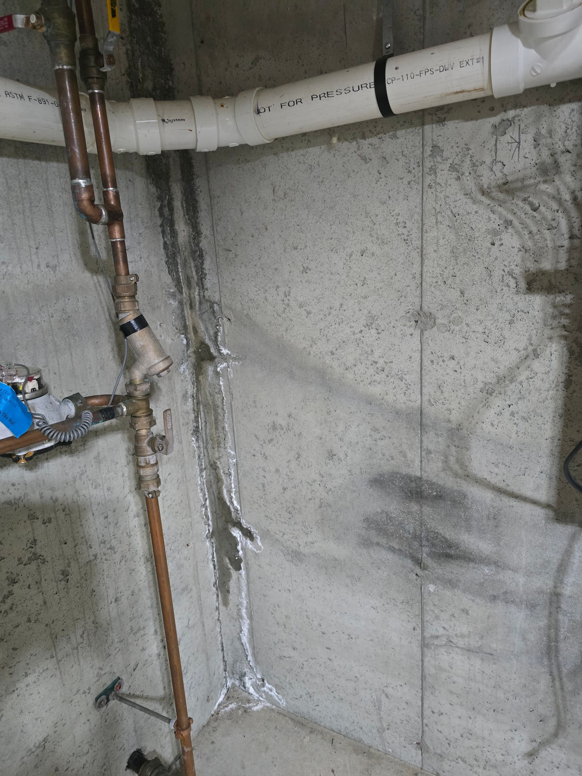 A concrete corner in a basement shows a patched vertical crack with copper plumbing and white PVC pipes above.