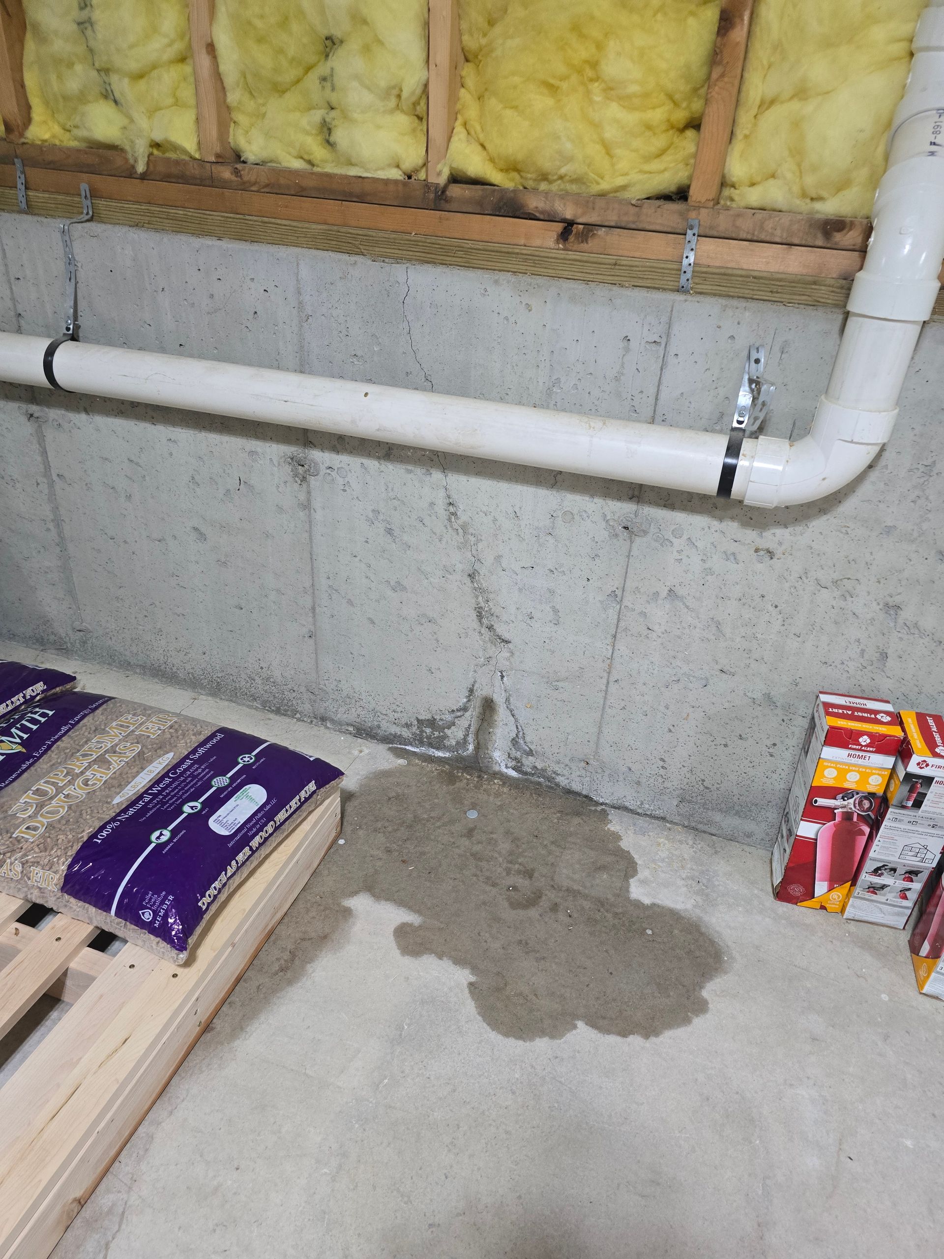 A wet patch on a concrete basement floor beneath a vertical crack in the foundation wall near a white PVC pipe.