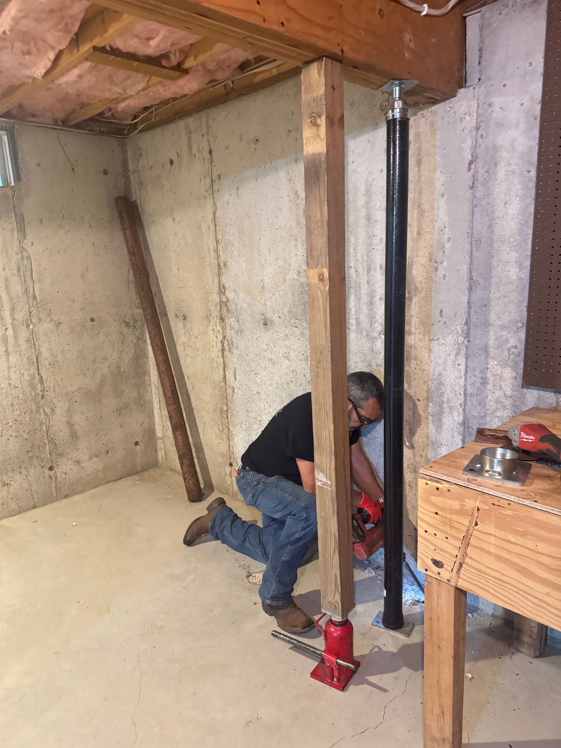 A person in a basement uses a hydraulic jack to lift a wooden post supporting a floor joist next to a steel column.