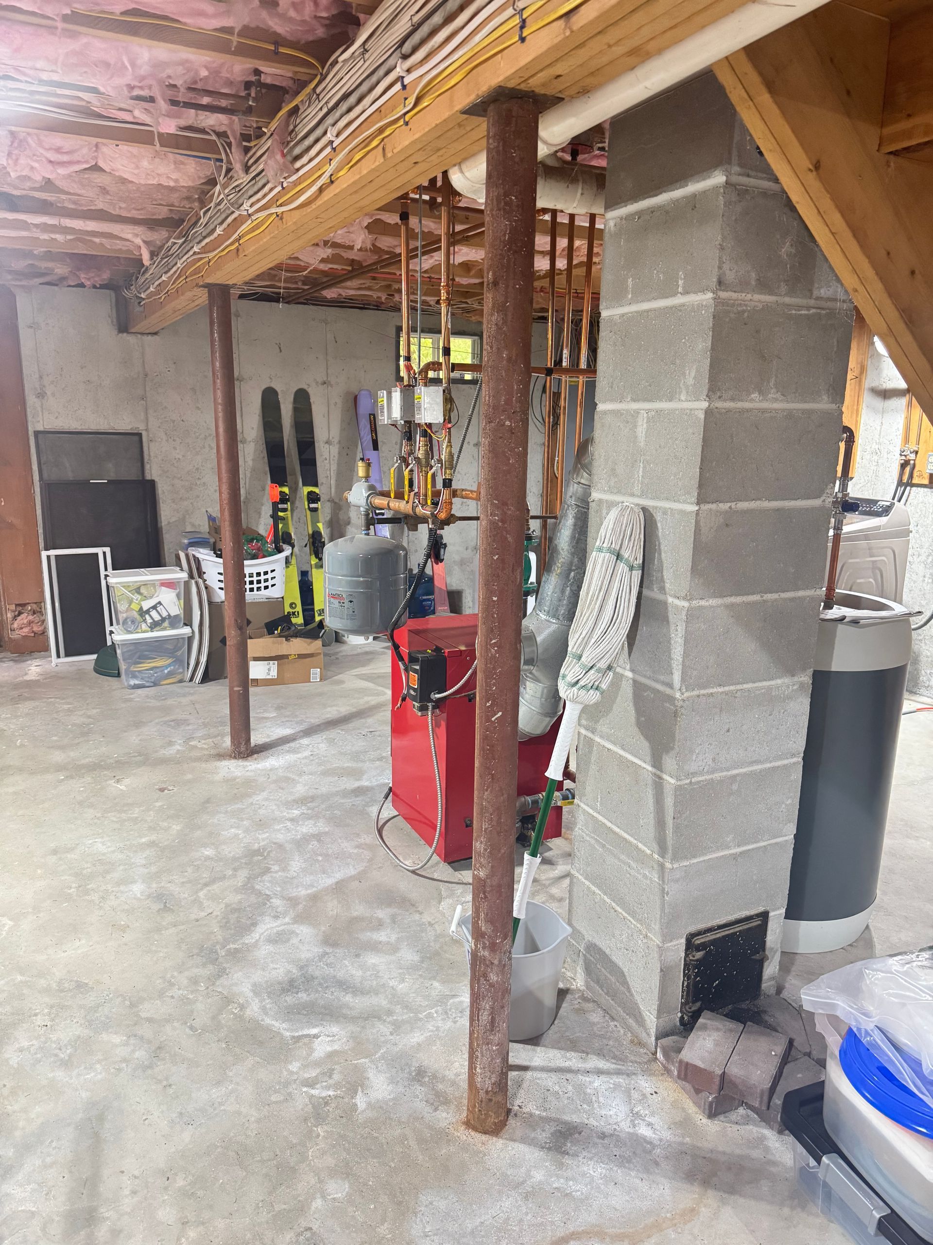 A cluttered basement with exposed ceiling insulation, metal support columns, a red furnace, and a concrete block pillar.