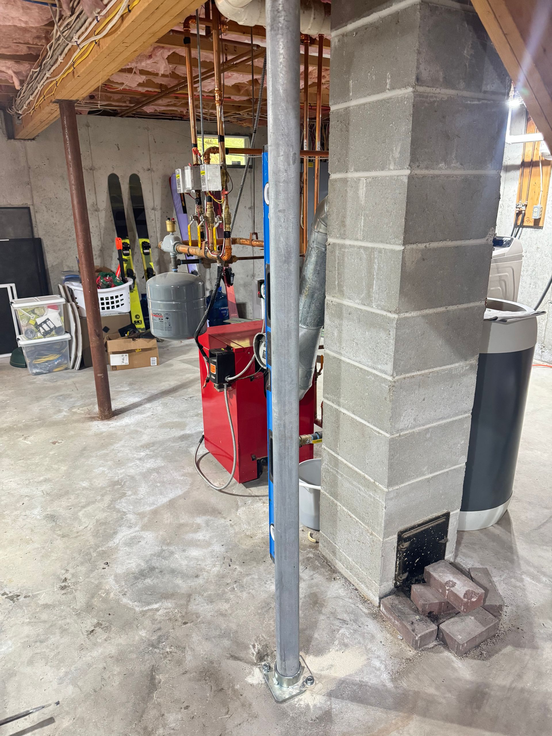 A basement with a metal support column, a concrete chimney, plumbing pipes, and a red boiler in the background.