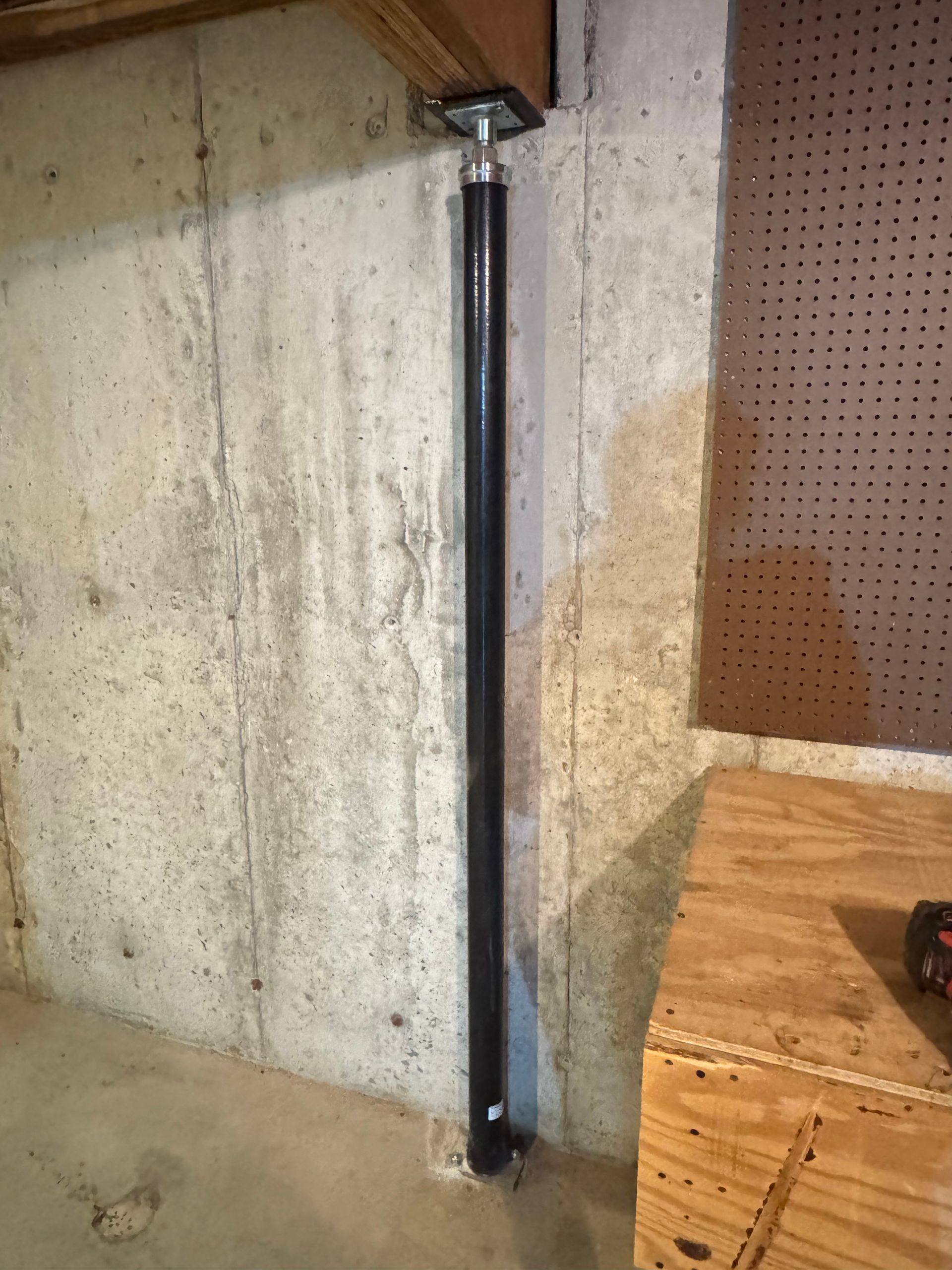 A black metal jack post supporting a wooden beam against a basement concrete wall.