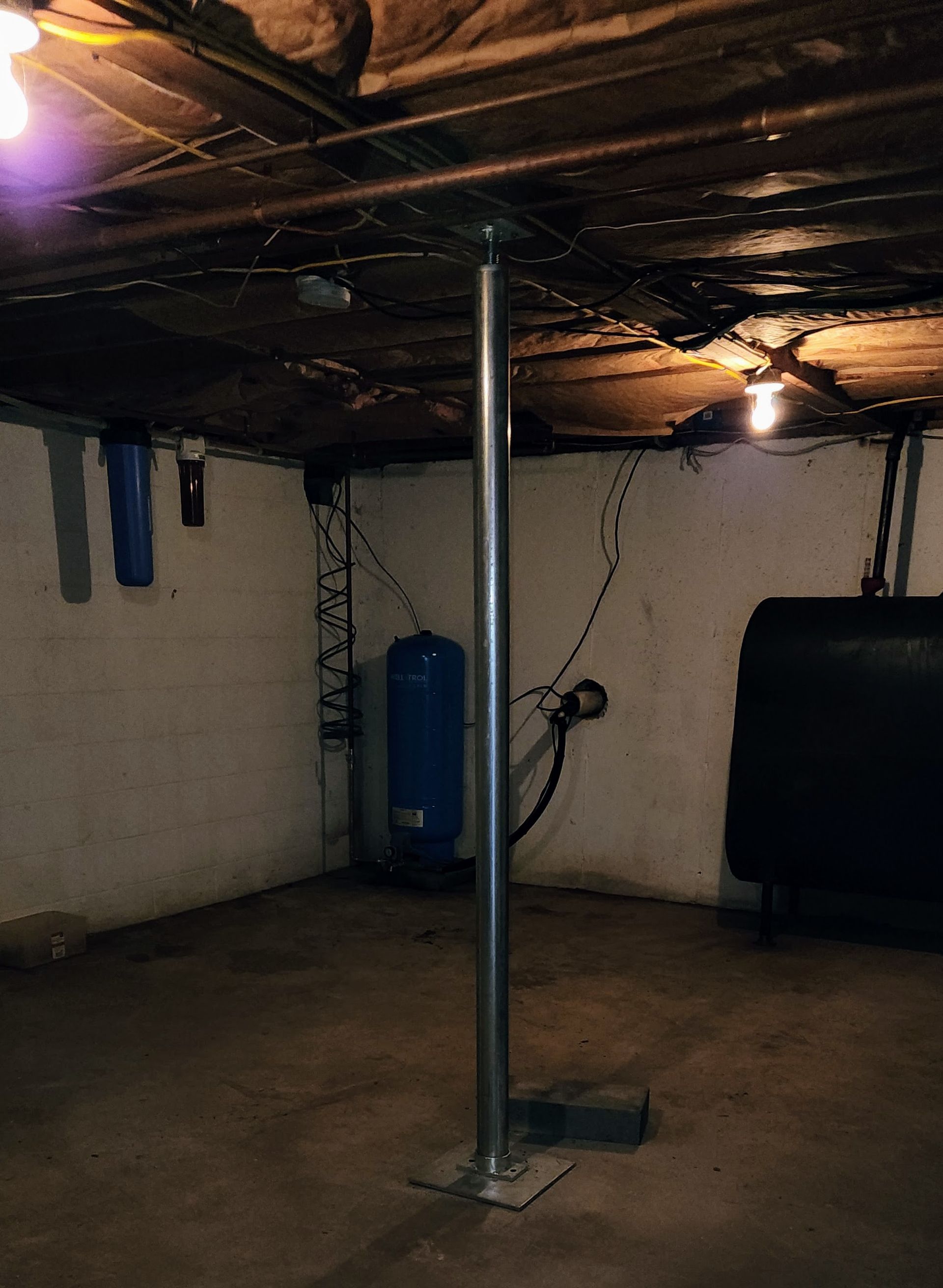 A vertical metal support post installed in a dimly lit unfinished basement with concrete walls and floor.