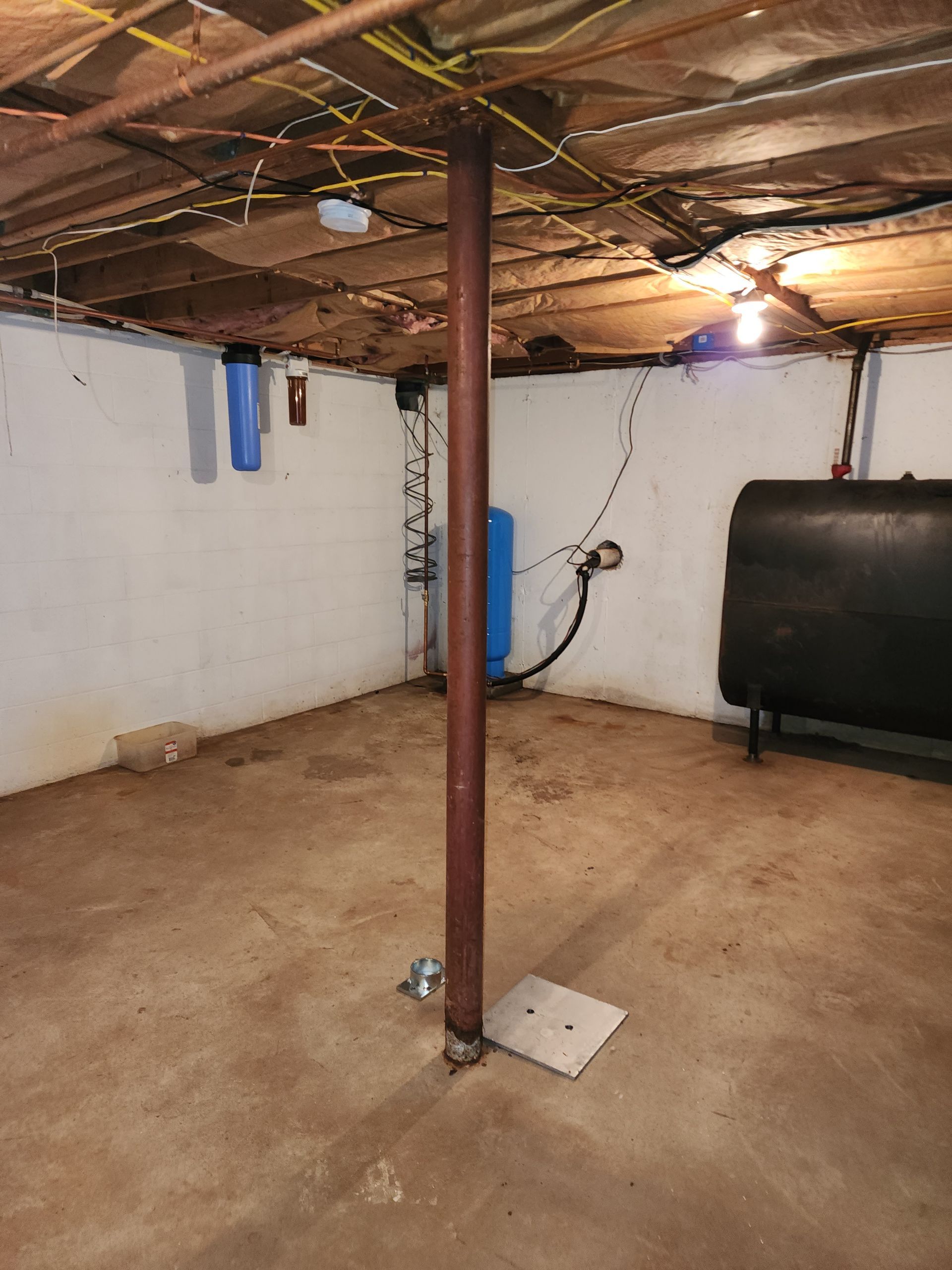 A basement with concrete walls and floor, featuring a central metal support column, a water tank, and an oil tank.