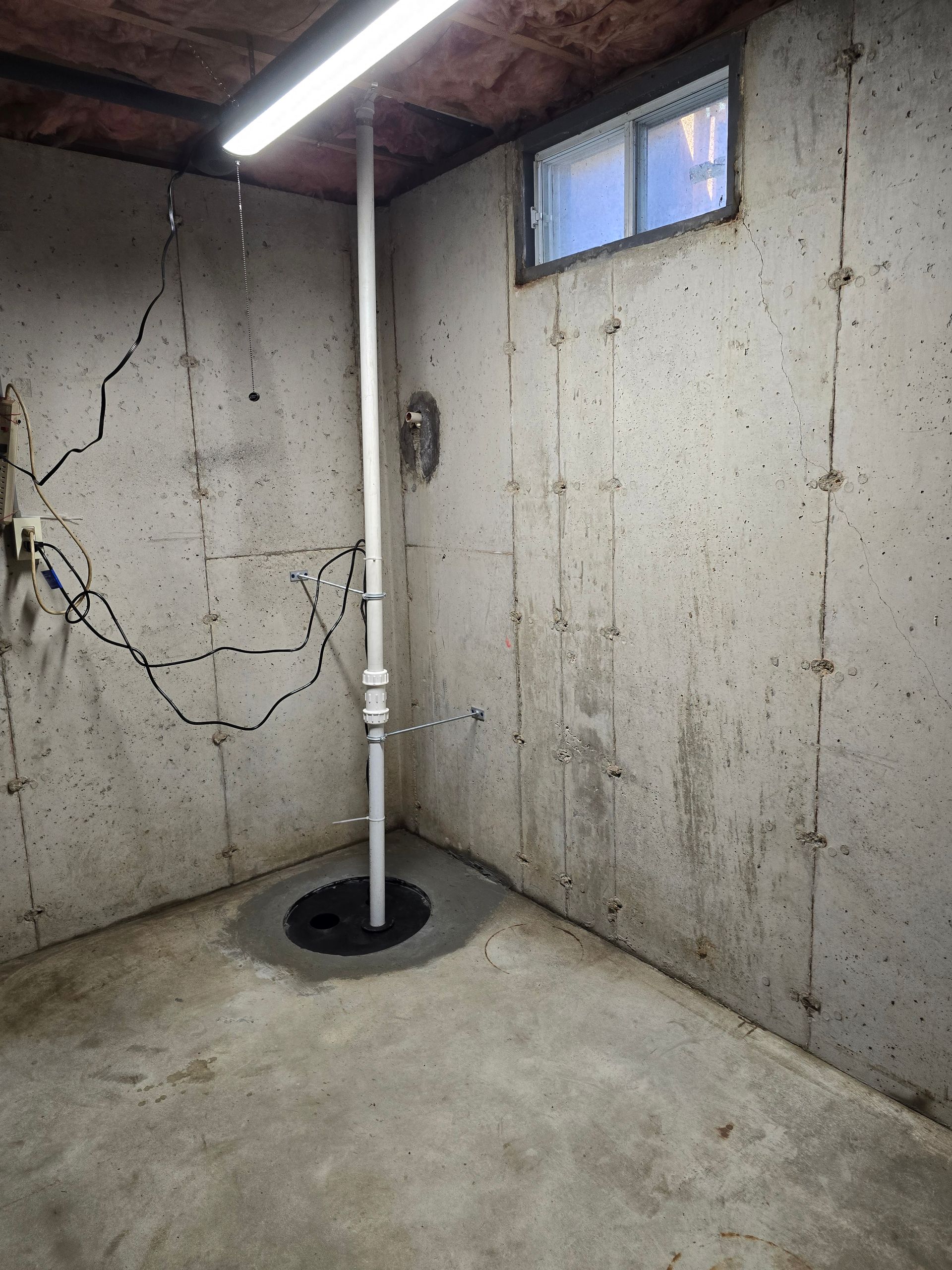 Basement corner with a sump pump and discharge pipe installed in the concrete floor near a small window.