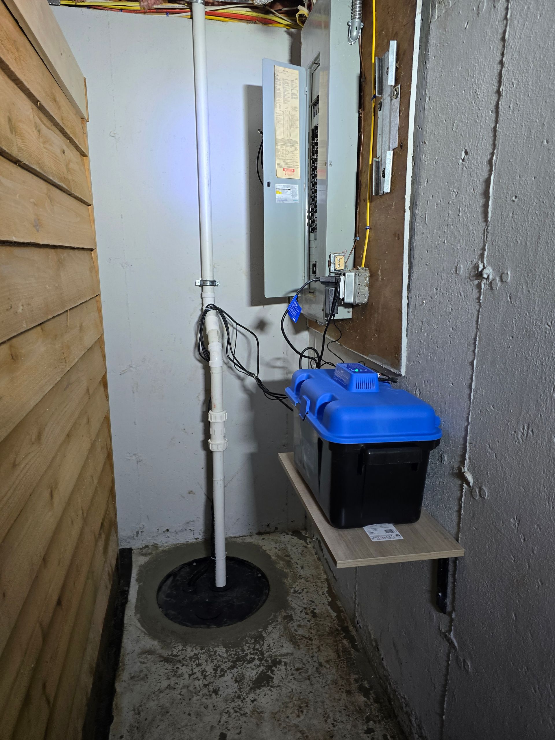 A sump pump with a white vertical discharge pipe inside a basement, next to a black and blue battery backup system.