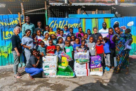 Large group posing outdoors with donated food and supplies in front of a colorful wall.