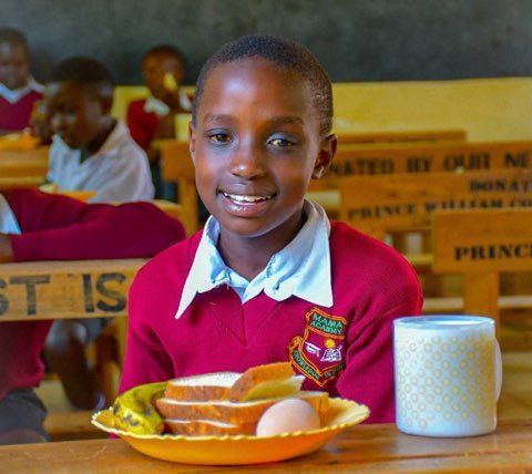 Student in a red sweater smiles at a desk with a tray of food and a white mug in a classroom.