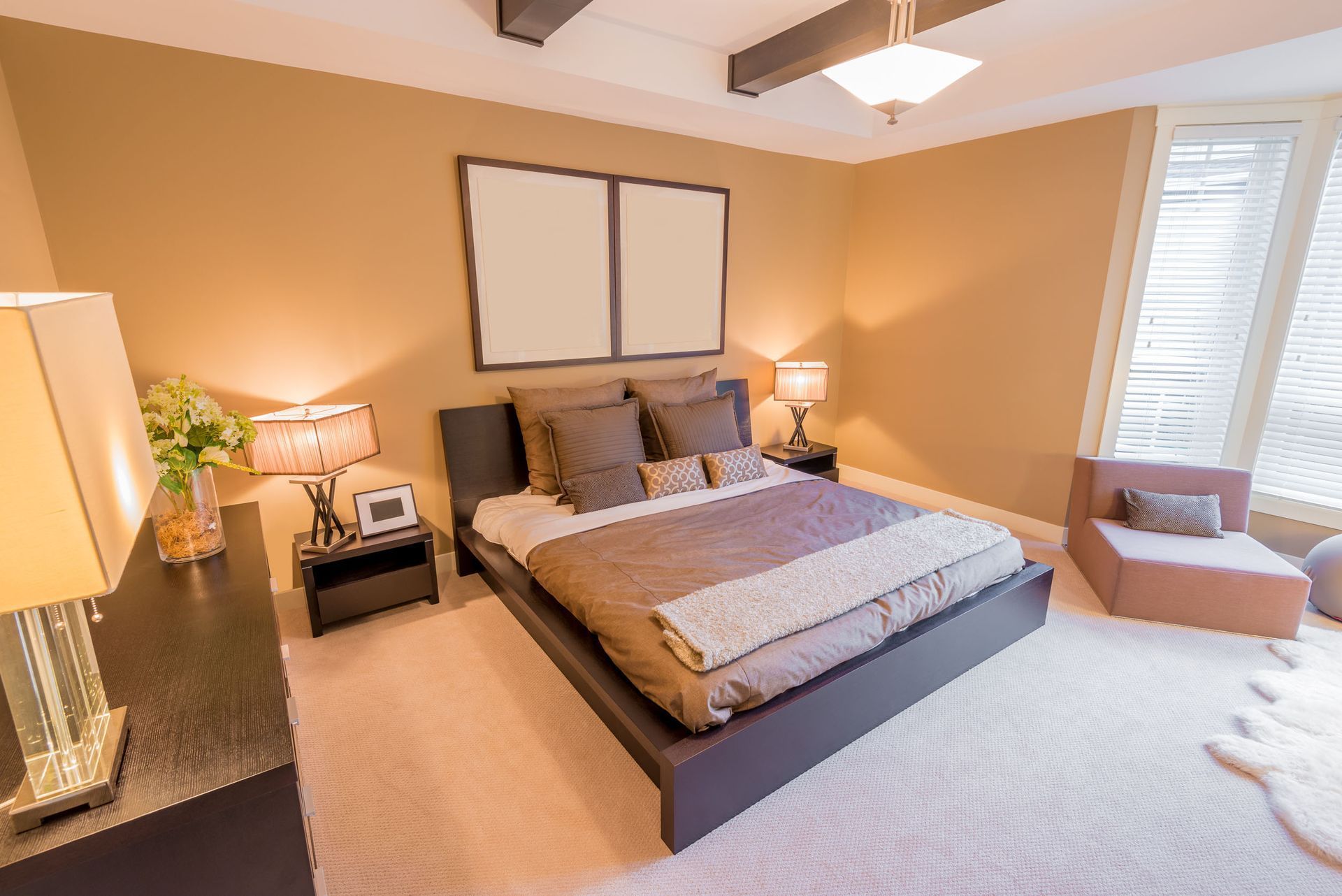 Bedroom with a bed, nightstands, chair, and a window with blinds; tan walls and carpet.