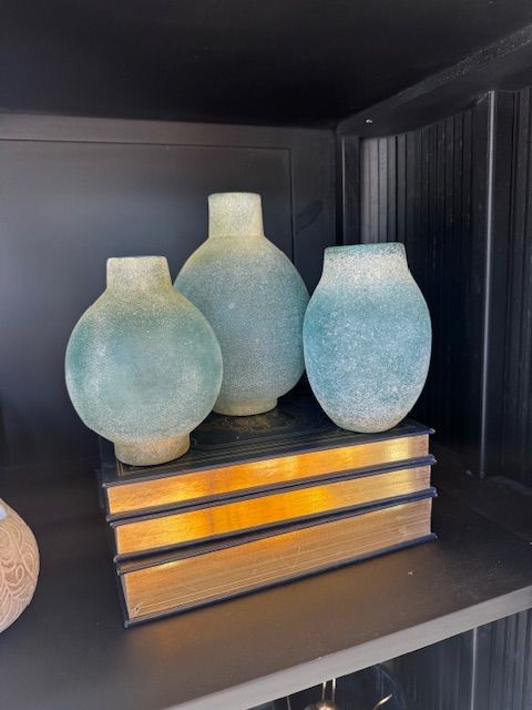 Three blue frosted vases on gold-edged books inside a dark shelf.