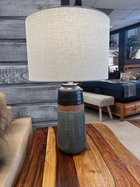 Lamp with a gray, blue, and copper base, topped by a beige lampshade, on a wood table.