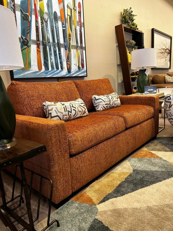 Orange textured couch with patterned pillows on a multicolored geometric rug, next to a lamp and painting.