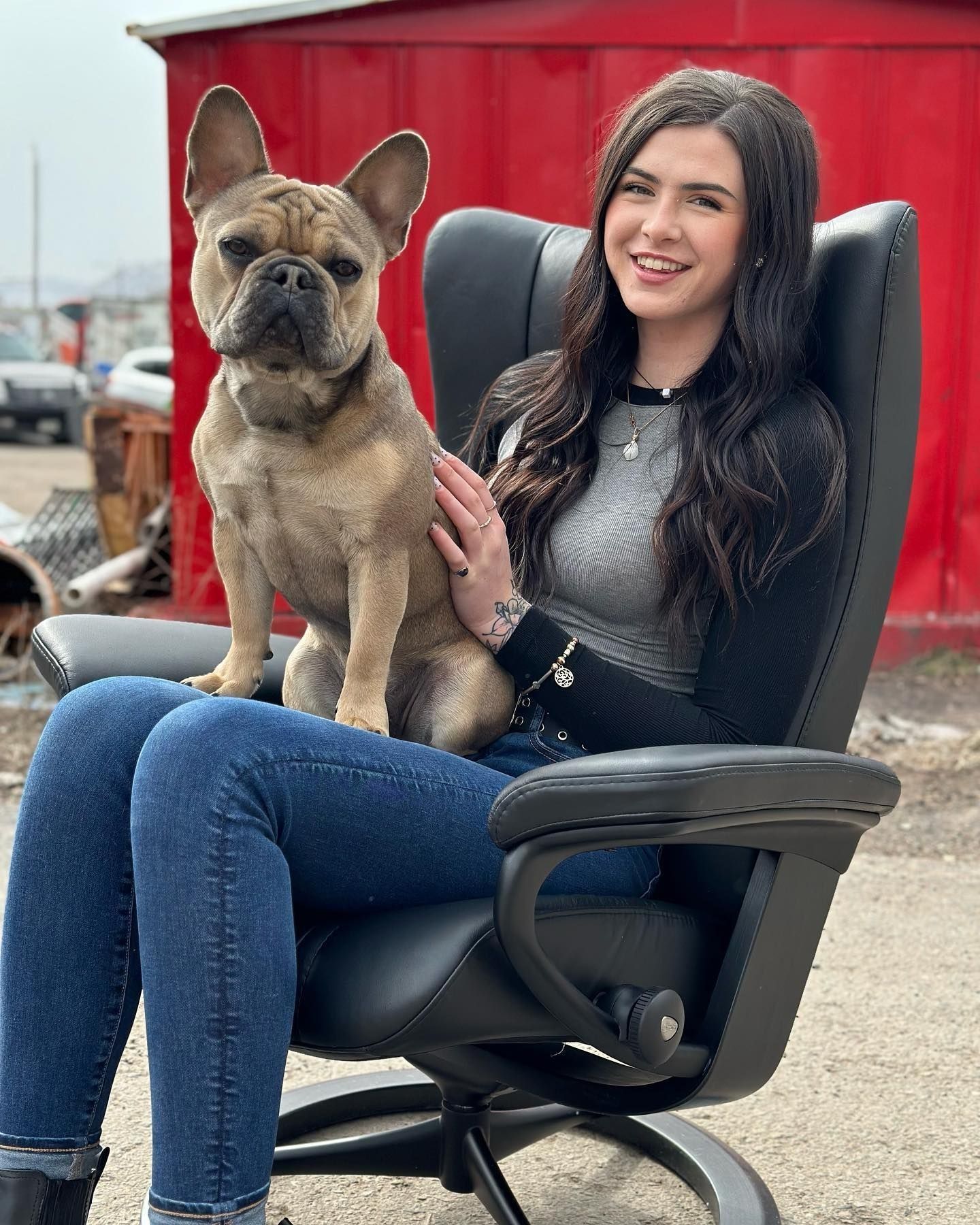 Woman and French bulldog seated in a black office chair. Both smiling, set against a red building and outdoors.