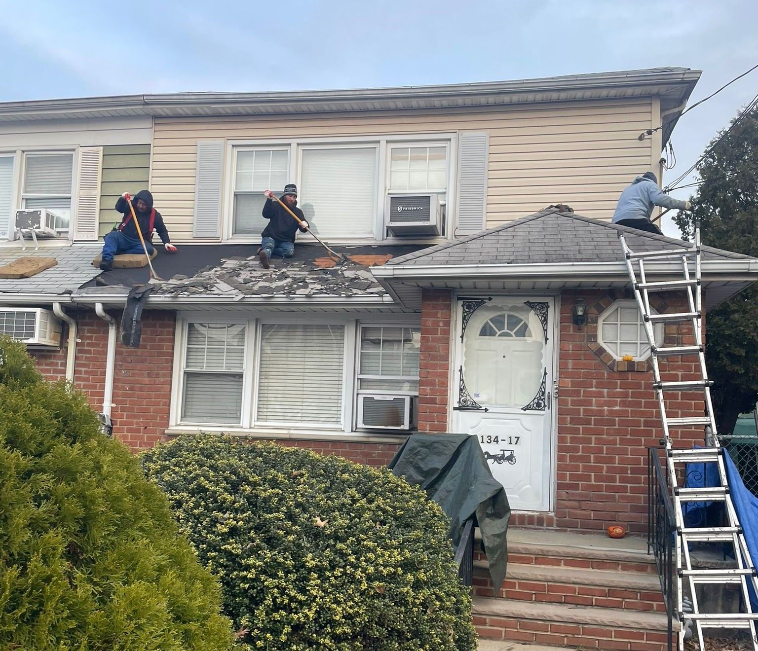 Three people on a roof removing shingles from a two-story brick building. One person on a ladder is working on the lights.