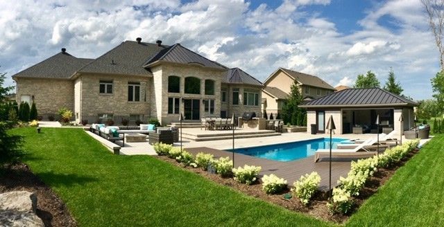 Luxury backyard with a pool, patio, and cabana, backing onto a large, light-colored brick house on a sunny day.