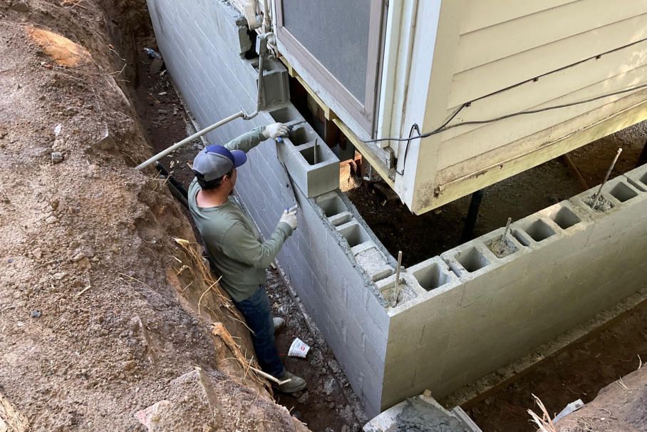 Man building cinder block foundation near a house. He’s wearing a blue cap, and the setting is outdoors with dirt.