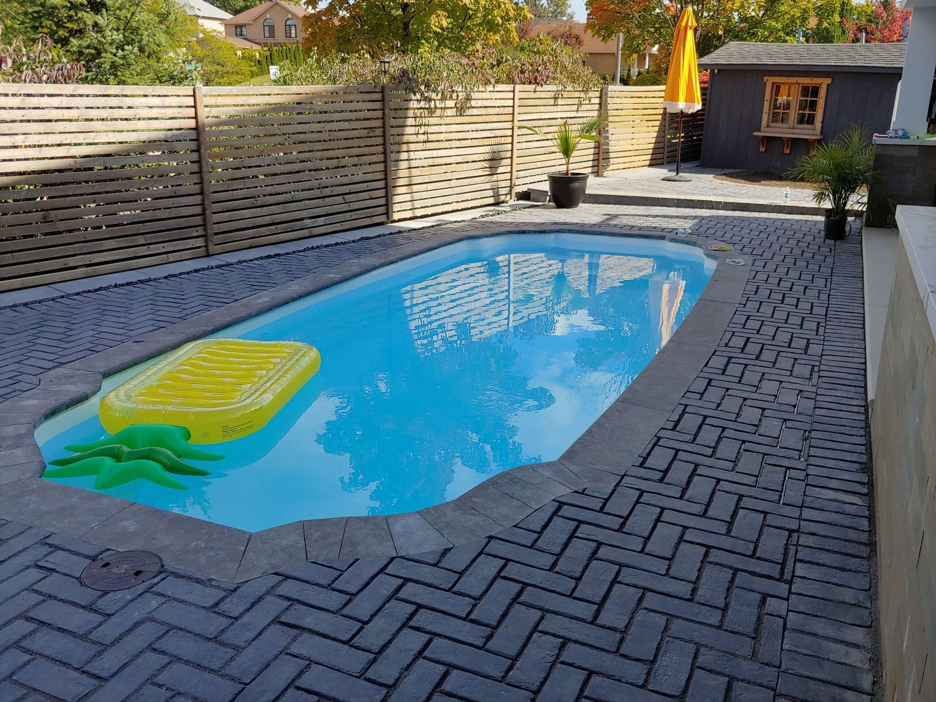 Rectangular backyard pool with blue water, surrounded by grey brick. A yellow pineapple float is inside.