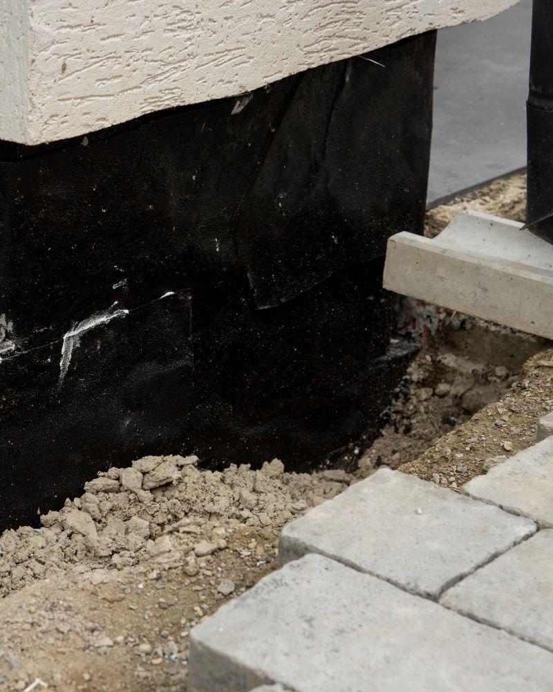 A close-up view of a building foundation coated in black waterproofing material next to a partially paved concrete walkway.