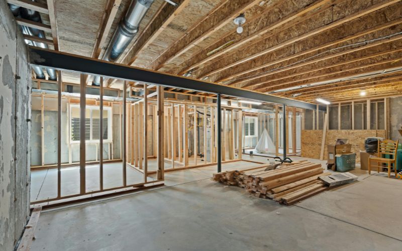 An unfinished basement with exposed wooden joists, concrete floor, stud-framed wall partitions, and a steel support beam.