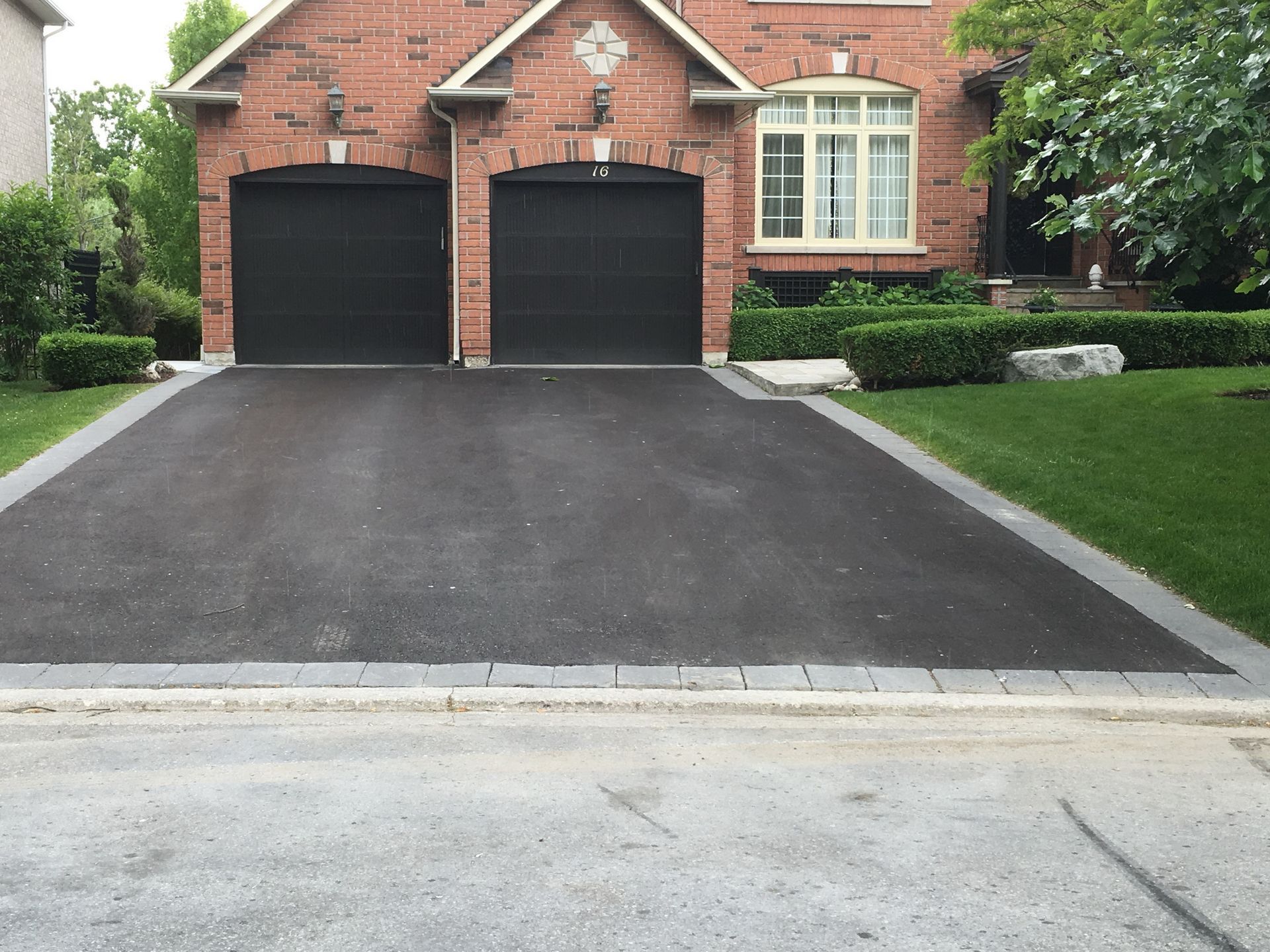 Asphalt driveway leading to a brick house with two black garage doors and trimmed hedges.