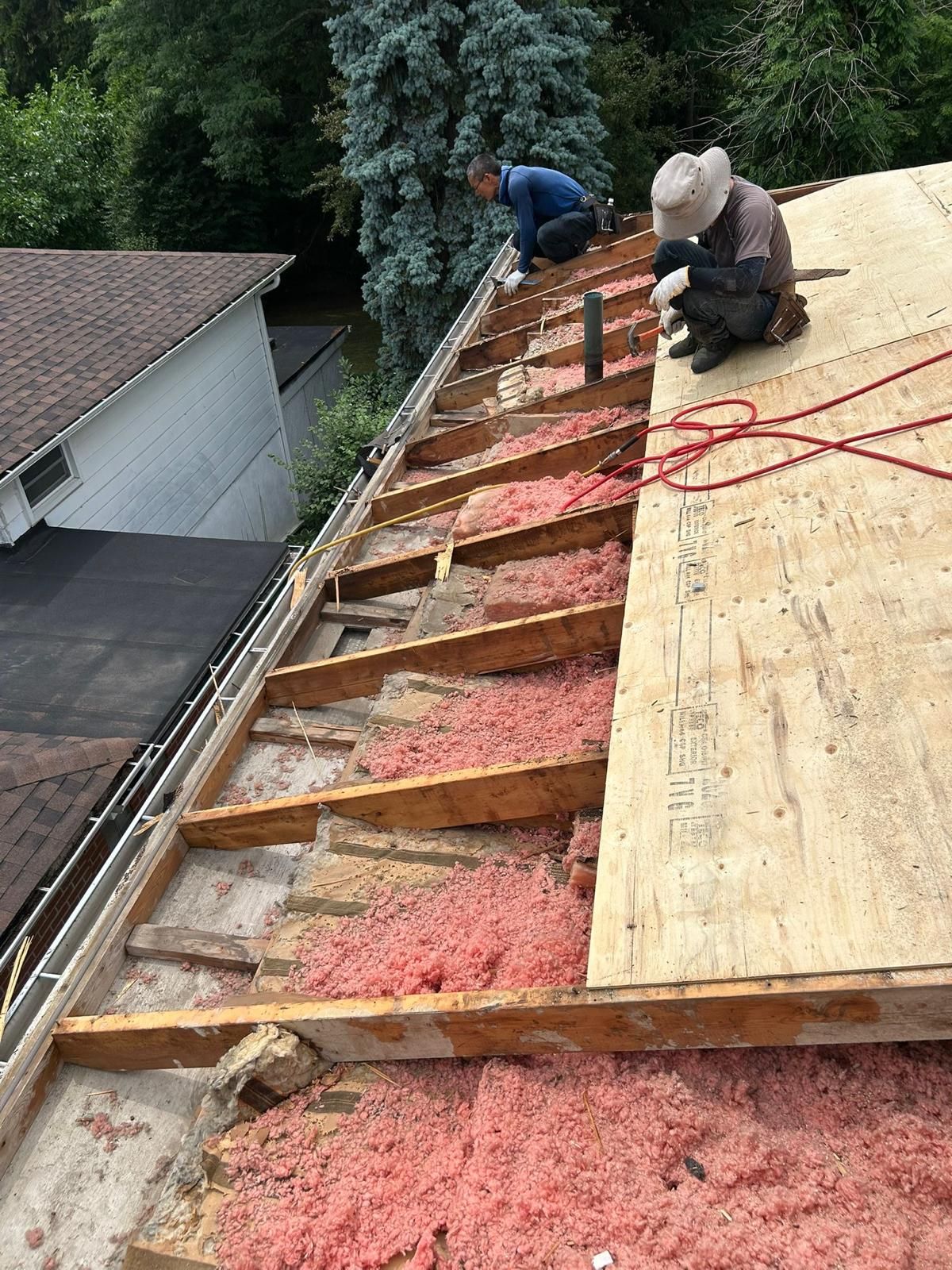 Two people on a roof replacing wood and insulation. Pink insulation is exposed.